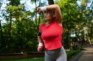 schön Athlet Frau, Läufer abwischen das Schweiß von ihr Gesicht mit Frottee Armband während Laufen im das Stadt Park, behalten ihr Körper fit. gesund und aktiv Lebensstil Konzept foto