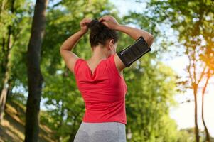 Rückseite Aussicht von ein Sportlerin tragen ein Smartphone Halter, binden Pferdeschwanz, bekommen bereit zum Lauf entlang das Stadt Park draussen foto