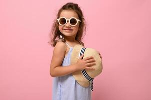 isoliert Porträt von schön bezaubernd 4 Jahre Kind Mädchen tragen Blau Sommer- Kleid und Sonnenbrille, posiert zu Kamera mit Stroh Hut im ihr Hände. Rosa Hintergrund mit Kopieren Raum foto