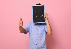 ein Schule Junge Abdeckungen seine Gesicht mit ein Tafel mit gezeichnet lächelnd Emoticons, ausdrücken Glück und zeigt an Daumen oben zu das Kamera. isoliert Über Rosa Hintergrund mit Kopieren Raum foto