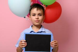 gut aussehend Junge mit leer Tafel im seine Hand Stehen gegen mehrfarbig Ballons auf Rosa Hintergrund mit Kopieren Raum foto