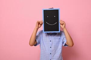 ein Schule Junge Abdeckungen seine Gesicht mit ein Tafel mit gezeichnet lächelnd Emoticons, ausdrücken Glück. isoliert Über Rosa Hintergrund mit Kopieren Raum foto