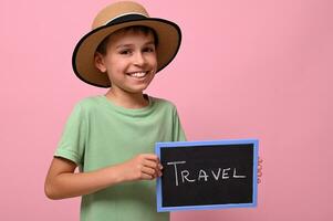 bezaubernd Junge halten ein Tafel mit Reise Beschriftung, lächelnd, suchen beim das Kamera. Rosa Hintergrund mit Kopieren Raum foto