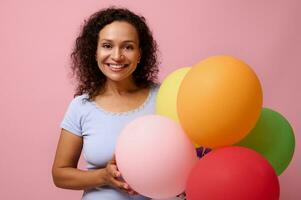 gewinnend attraktiv gemischt Rennen Frau mit ein charmant zahnig lächeln, hat glücklich Reaktion halten hell bunt Helium Luftballons , feiert Geburtstag, sieht aus beim Kamera, isoliert auf Rosa farbig Hintergrund foto