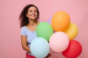 gewinnend gemischt Rennen Frau mit ein charmant zahnig lächeln, hat glücklich Reaktion halten hell bunt Helium Luftballons , feiert Geburtstag, sieht aus beim Kamera, isoliert auf hell Rosa farbig Hintergrund foto