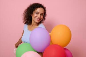 gewinnend schön gemischt Rennen Frau mit ein charmant zahnig lächeln, hat glücklich Reaktion halten hell bunt Helium Luftballons , feiert Geburtstag, sieht aus beim Kamera, isoliert auf Rosa farbig Hintergrund foto