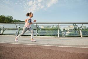 Seite voll Länge Porträt von ein entschlossen aktiv sportlich Frau Laufen schnell entlang das modern Glas Stadt Brücke Laufband. aktiv und gesund Lebensstil, draussen Herz, Gewicht Verlust, Laufen Konzept foto