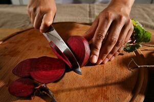 Nahansicht. Frau Koch Hände halten ein Küche Messer und Hacken Scheiben von frisch roh saisonal Rüben auf ein hölzern Schneiden Tafel foto