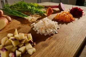 geschreddert Rübe, Karotte, Zwiebel und Würfel von Schnitt Kartoffel auf ein hölzern Tafel mit Hälften von Gemüse mit Grün Blätter und Wurzeln auf das Hintergrund. Nahansicht. Essen Hintergrund foto