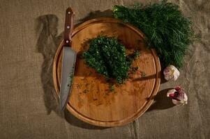 oben Aussicht von ein Bündel von frisch feucht Dill auf ein Leinen- Tischdecke und Knoblauch Nächster zu ein hölzern Schneiden Tafel mit ein Küche Messer und gehackt Dill. saisonal Grüns Essen Hintergrund. eben legen. foto