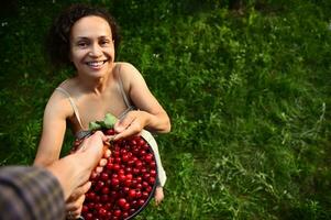 Ernte Kirschen im das Garten von ein Land Haus. ein Mann auf ein Trittleiter dient ein Eimer von Kirschen zu ein charmant lächelnd afrikanisch Frau im ein Leinen- Kleid foto