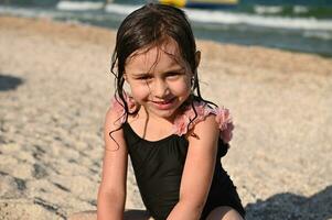 Kopfschuss. bezaubernd ziemlich Baby Mädchen mit nass Haar und Badeanzug, Sitzung auf das sandig Strand auf das Meer Hintergrund. Kinder Emotionen, Glück. Sommer- Urlaub Konzept. süß Mädchen genießen Sommer- Urlaube foto