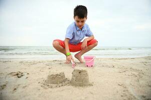 Glücklich, gut aussehend und bezaubernd Junge, Kind, Kleinkind Füllung Spielzeug Eimer mit Sand zum Gebäude Sandburgen beim das Strand. Natur, Seelandschaft Hintergrund. Sommer- Freizeit Aktivitäten. foto