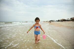 ziemlich wenig Mädchen hält ein Rosa Spielzeug Eimer im ihr Hand und Spaziergänge entlang das Meeresküste, genießen Sonnenbaden, draußen Erholung. Konzepte von glücklich und sorglos Sommer- Urlaube beim das Strand foto