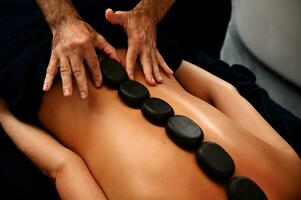 Nahansicht von Masseur Verlegung heiß Steine entlang Rücken von jung Frau und tun ayurvedisch heiß Stein Massage im modern Spa Salon foto