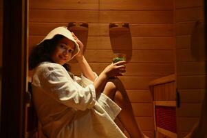 ein schön Frau dämpfen im das Sauna mit ein Glas von gesund trinken im ihr Hand. Haut und Körper Pflege Konzepte. Gesundheit Pflege und Schönheit Behandlung im Spa Center foto