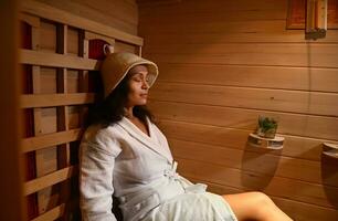 heiter Frau im Waffel Bademantel und Sauna Hut entspannend im Infrarot Sauna foto
