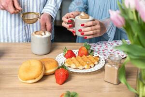 ein mans und ein Frau Hände im Licht Blau Kleider mit rot Nägel. ein Mann Anzeigen Zimt im ein grau Tasse mit Kaffee Latté. das knusprig Waffel, rot Erdbeeren auf ein Weiß Teller und Donuts. foto