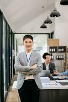 jung attraktiv asiatisch männlich Büro Arbeiter Geschäft Anzüge lächelnd beim Kamera mit Arbeiten Notizblock, Tablette und Laptop Unterlagen im Büro foto