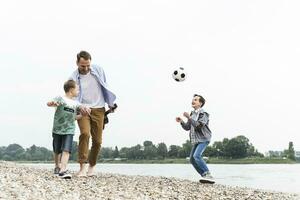 glücklich Vater mit zwei Söhne und Fußball Gehen beim das Flussufer foto