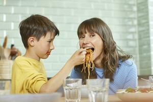 Sohn Fütterung Mutter mit Spaghetti im das Küche foto