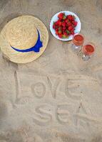 Stroh Hut, Wein und ein Teller von Erdbeeren auf ein sandig Strandinschrift Liebe das Meer foto