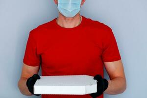 Lieferung Mann Mitarbeiter im rot Deckel leer T-Shirt Uniform Gesicht Maske Handschuhe halt Karton Box Pizza auf grau Hintergrund Bedienung Quarantäne Pandemie Coronavirus Virus 2019-ncov Konzept. foto