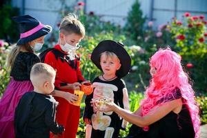 Kinder Trick oder behandeln im Halloween Kostüm und medizinisch Maske. ein wenig Junge, Mädchen und Baby im Anzüge während das Coronavirus Pandemie erhalten Süßigkeiten von ein Frau . foto