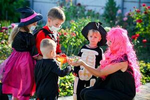 Kinder Trick oder behandeln im Halloween Kostüm und medizinisch Maske. ein wenig Junge, Mädchen und Baby im Anzüge während das Coronavirus Pandemie erhalten Süßigkeiten von ein Frau. foto