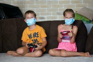 wenig Mädchen und Junge abspielen medizinisch Maske Spiel Konsole während Sitzung auf Sofa beim heim. foto