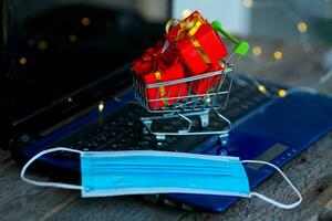 klein Korb mit Kisten von Weihnachten Geschenke und ein medizinisch Maske auf das Hintergrund von ein Laptop. online Einkaufen. Boxen Tag Konzept im das covid-19 Pandemie. foto