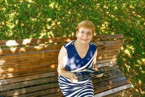 Senior Frau lächelnd mit ein Tablette im ihr Hände auf ein Park Bank auf ein sonnig Tag, oben Aussicht foto