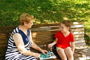 Oma und Enkelin stellen zusammen ein Puzzle im das Park auf ein Bank auf ein sonnig Tag. foto