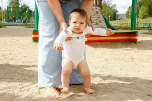 Baby nimmt zuerst Schritte mit Mamas Hilfe auf das Spielplatz foto