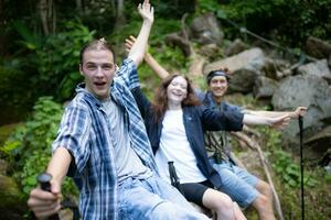 Gruppe von freunde mit Rucksäcke und Stöcke Sitzung auf ein gefallen Baum, nehmen ein brechen während das Wanderung foto