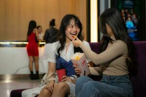 zwei jung asiatisch Frauen Essen Popcorn und warten zum Aufpassen Film beim Vorderseite von Kino foto