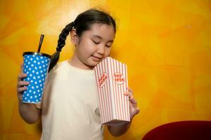 asiatisch Kind Mädchen Essen Popcorn und Trinken Limonade auf Gelb Mauer Hintergrund, Kino Konzept, foto