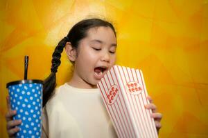 asiatisch Kind Mädchen Essen Popcorn und Trinken Limonade auf Gelb Mauer Hintergrund, Kino Konzept, foto