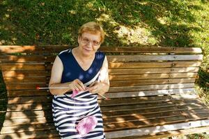 Senior Frau strickt im das Park auf ein Bank auf ein sonnig Tag foto