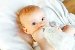 süß wenig Neugeborene Mädchen Trinken Milch von Flasche und suchen beim Kamera auf Weiß Hintergrund. Säugling Baby saugen Essen Milch Ernährung Lügen Nieder auf Krippe Bett beim heim. Mutterschaft glücklich Kind Konzept. foto