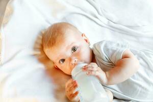 süß wenig Neugeborene Mädchen Trinken Milch von Flasche und suchen beim Kamera auf Weiß Hintergrund. Säugling Baby saugen Essen Milch Ernährung Lügen Nieder auf Krippe Bett beim heim. Mutterschaft glücklich Kind Konzept. foto