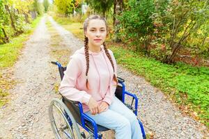 junge glückliche Frau mit Handicap im Rollstuhl auf der Straße im Krankenhauspark, die auf Patientendienste wartet. gelähmtes Mädchen im Rollstuhl für Behinderte im Freien in der Natur. Rehabilitationskonzept. foto