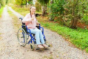 junge glückliche Frau mit Handicap im Rollstuhl auf der Straße im Krankenhauspark, die auf Patientendienste wartet. gelähmtes Mädchen im Rollstuhl für Behinderte im Freien in der Natur. Rehabilitationskonzept. foto