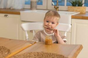 glücklich Familie beim heim. Baby Junge Fütterung selbst im Küche. wenig Junge mit unordentlich komisch Gesicht isst gesund Lebensmittel. Kind lernt Essen durch selbst halten Löffel. selbst füttern. foto