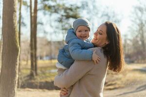 glücklich Familie draussen. Mutter Umarmen ihr Kind draussen. Mama Heben im Luft wenig Kleinkind Kind Sohn. Frau und wenig Baby Junge ruhen Gehen im Park. Mutter Umarmungen Baby mit Liebe Pflege. foto