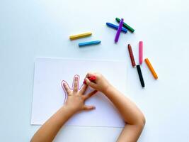 Kinder Hände zeichnen ihr Hand mit Wachs Buntstifte auf Weiß Papier, oben Sicht. foto