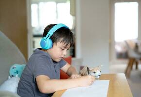 glücklich Kind tragen etwas Kopfhörer und Hören zu Musik- während Zeichnung auf Papier, Innen- Porträt durch süß Junge genießen ein kreativ Aktivität beim Zuhause auf ein Wochenende. ein Kind ist tun Hausaufgaben foto