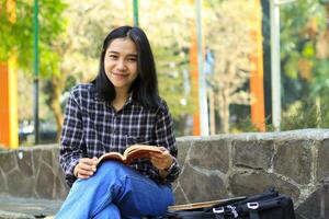 aufgeregt asiatisch schön jung Frau Hochschule Schüler lächelnd mit glücklich Gesicht lesen ein Buch draussen, Campus Universität Konzept foto