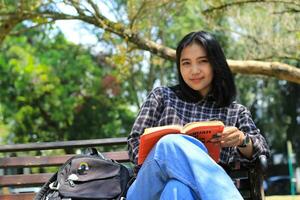 glücklich jung asiatisch Frau Hochschule Schüler mit lächelnd Gesicht genießen lesen ein Buch zum Prüfung im das Park foto