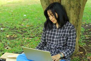 Porträt von konzentriert jung asiatisch Frau Surfen Internet mit Laptop zu Arbeiten aus der Ferne, lesen Buch, und Schreiben im Notizbuch. attraktiv asiatisch Schüler Studie im draußen foto